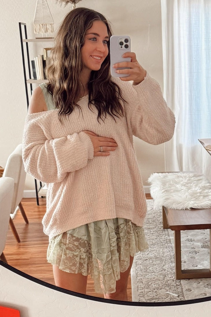 woman wearing a light pink oversized maternity sweater and a green lace dress, holding her baby bump in the mirror selfie