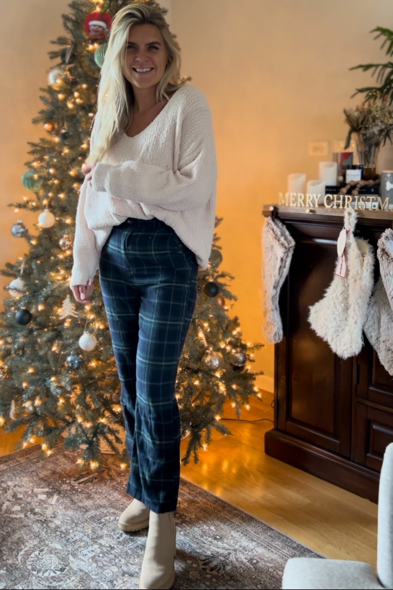 woman wearing a cream oversized sweater and dark green plaid flared pants, standing by a decorated Christmas tree