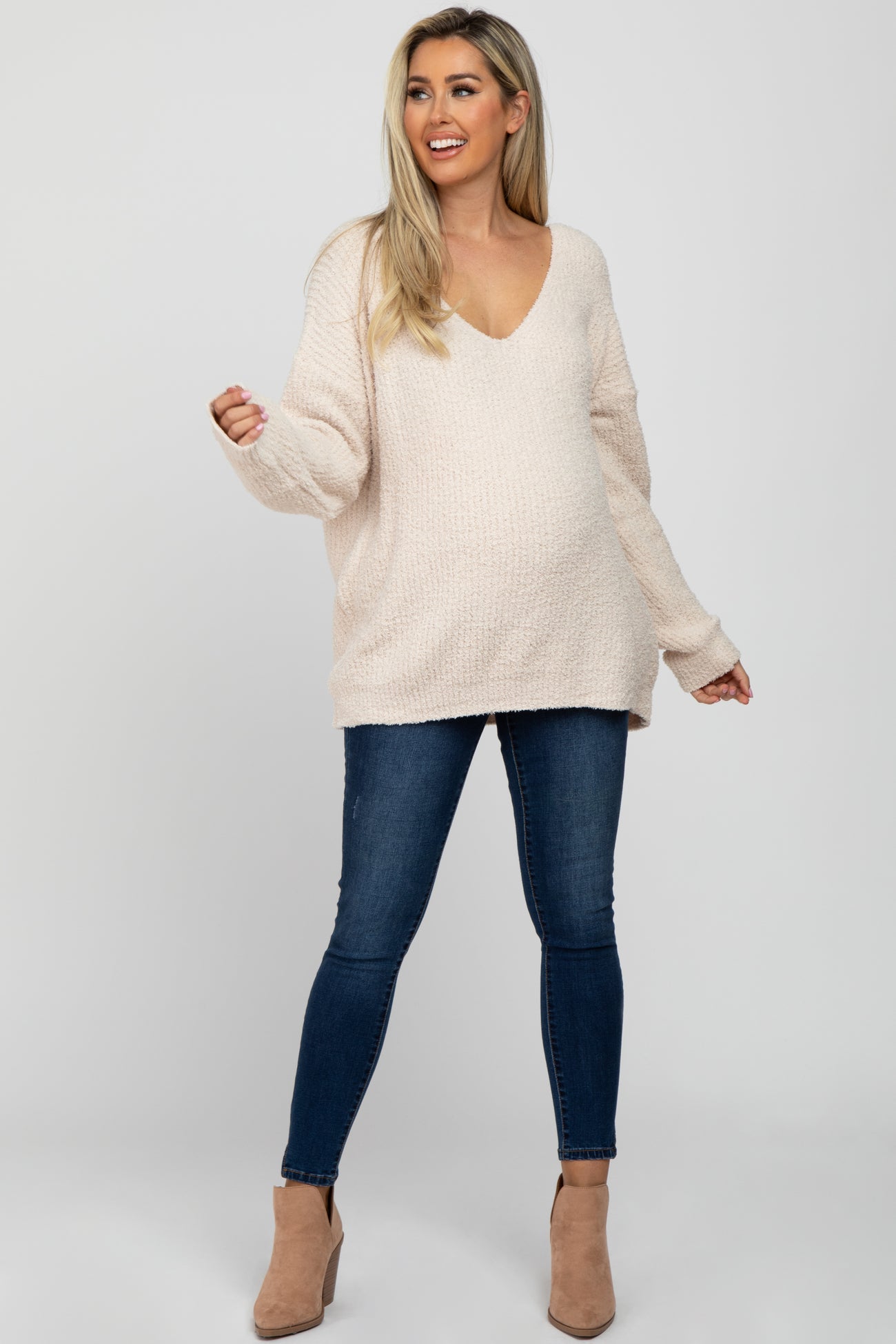 woman wearing a light beige oversized maternity sweater with a v-neck and distressed blue maternity jeans, showing her baby bump
