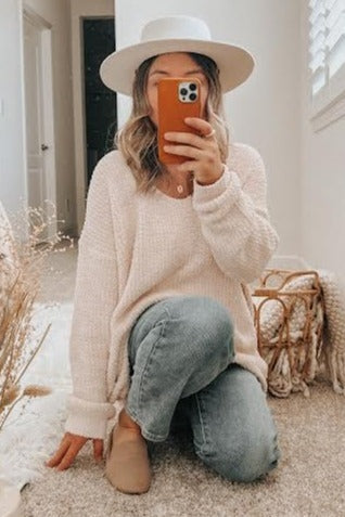 woman wearing a light pink oversized sweater, blue jeans, and a wide-brimmed hat, kneeling on a carpeted floor