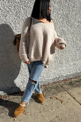 woman wearing a cozy beige oversized sweater, distressed jeans, and tan slip-on shoes, standing against a textured wall