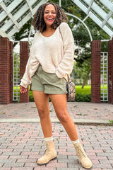 woman wearing a cream sweater and olive green shorts, styled with beige boots, showcasing her baby bump in maternity wear
