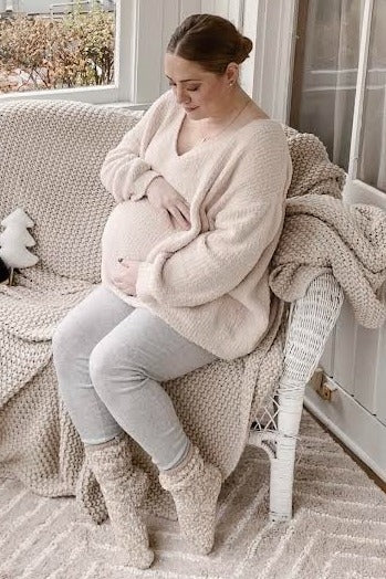 woman wearing a soft beige maternity sweater and gray leggings, cradling her baby bump while sitting on a cozy sofa