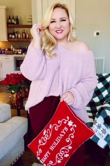 woman wearing a light pink off-shoulder sweater and holding a red decorative pillow with "happy holidays" written on it