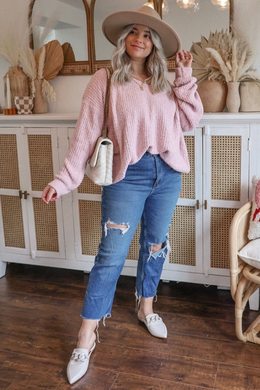 woman wearing a pink oversized sweater, distressed blue jeans, and white loafers, posing with a cream handbag