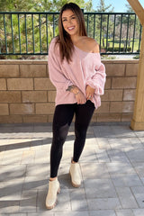 woman wearing a pink oversized sweater and black leggings, standing outdoors with a smile and hands on her hips