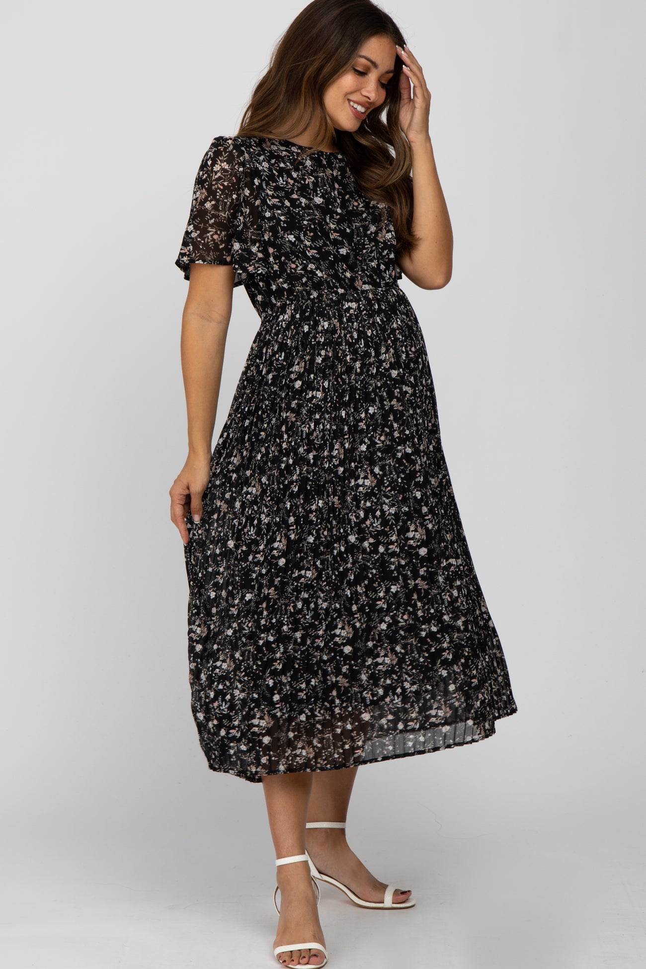 smiling pregnant woman wearing a black floral pleated maternity midi dress and white heels