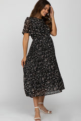 smiling pregnant woman wearing a black floral pleated maternity midi dress and white heels