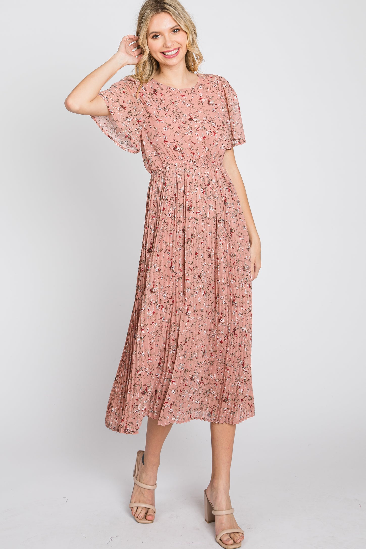 woman wearing a pink floral maxi dress with short sleeves and pleated skirt, posing with one hand in her hair