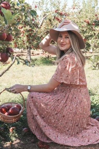 woman wearing a pink floral dress and wide-brimmed hat, sitting in an orchard and holding a basket of apples