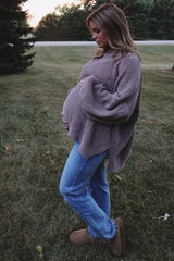 woman wearing a taupe oversized maternity sweater and blue jeans, cradling her baby bump in an outdoor setting