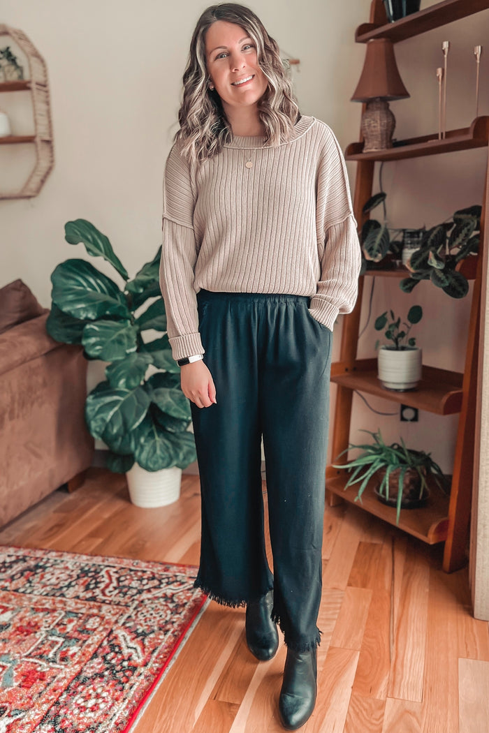 woman wearing a beige ribbed sweater and black wide-leg pants with frayed hems, standing in a cozy indoor setting
