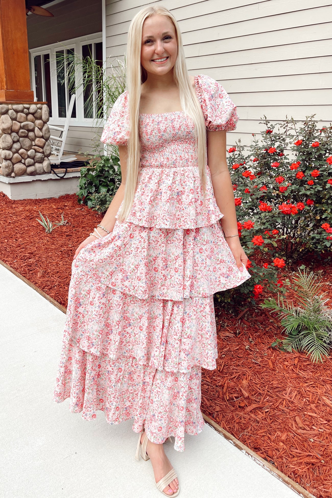 woman wearing a pink floral maxi dress with puff sleeves and tiered ruffles, standing outdoors with flowers in the background