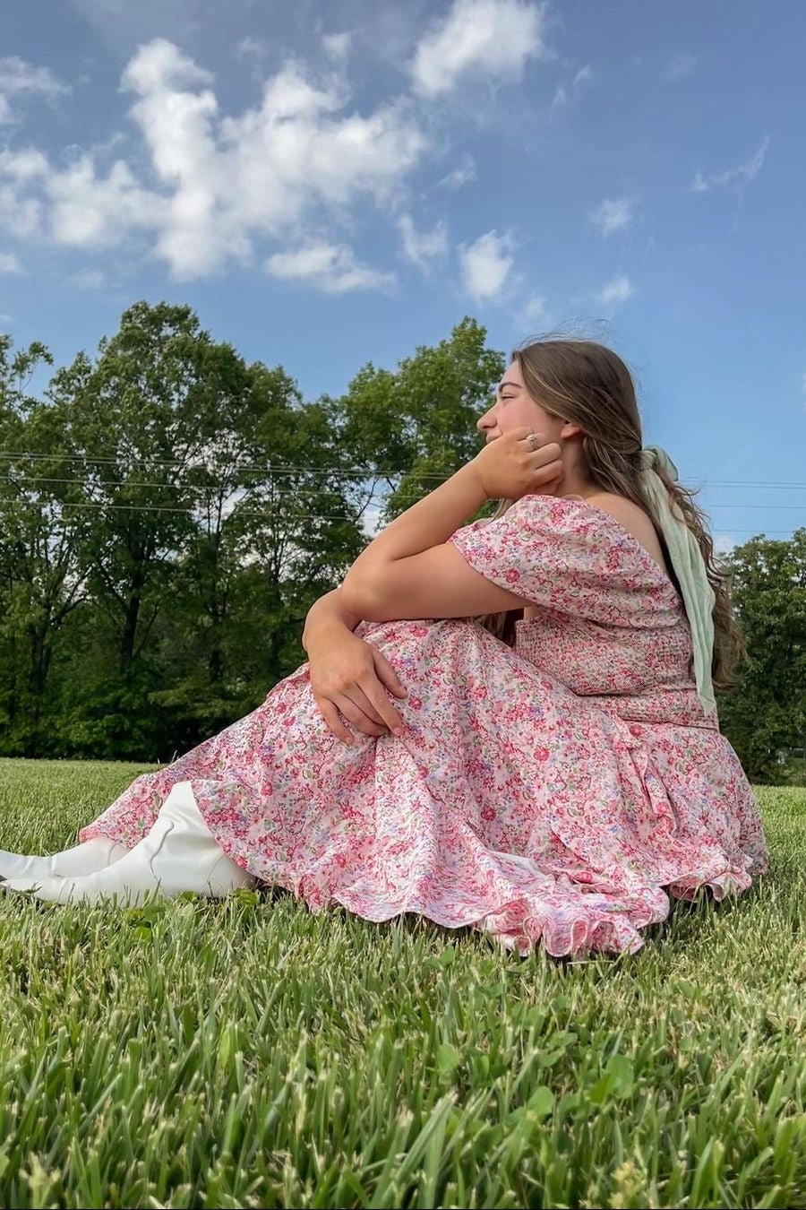 woman sitting on grass wearing a pink floral maxi dress and white knee-high boots, with her hair styled in loose waves