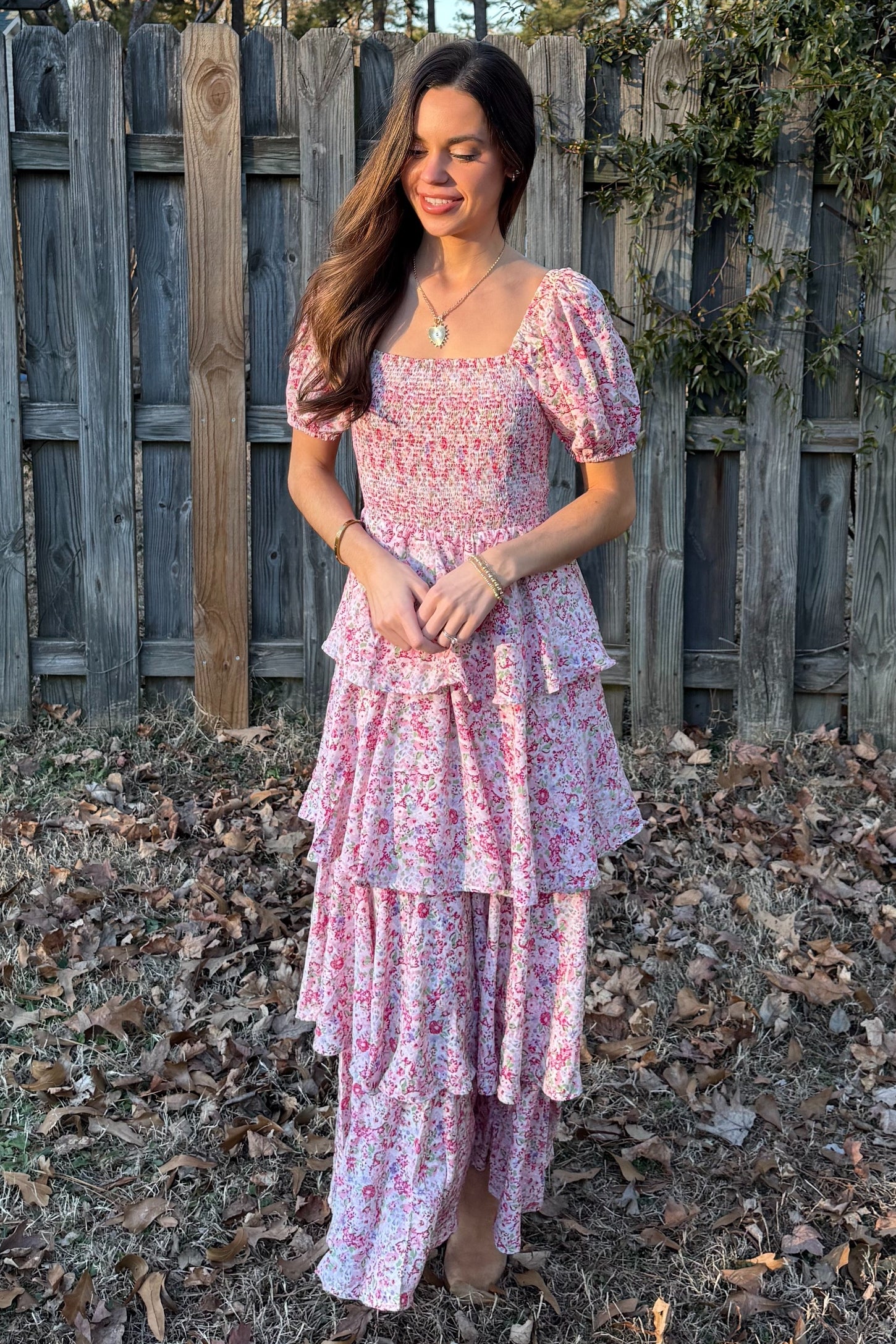 woman wearing a pink floral tiered maxi dress with puff sleeves, standing outdoors against a wooden fence