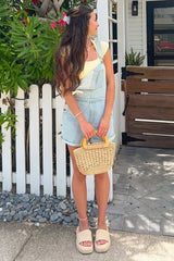 woman wearing a light yellow fitted t-shirt and light denim overalls, holding a woven handbag and wearing platform sandals