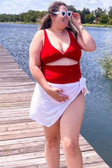 woman wearing a red cut-out one-piece swimsuit and white sarong, walking by the water, smiling and wearing heart-shaped sunglasses
