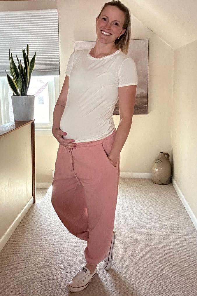 woman wearing a white short sleeve maternity top and pink joggers, showing her baby bump while posing in a sunlit room