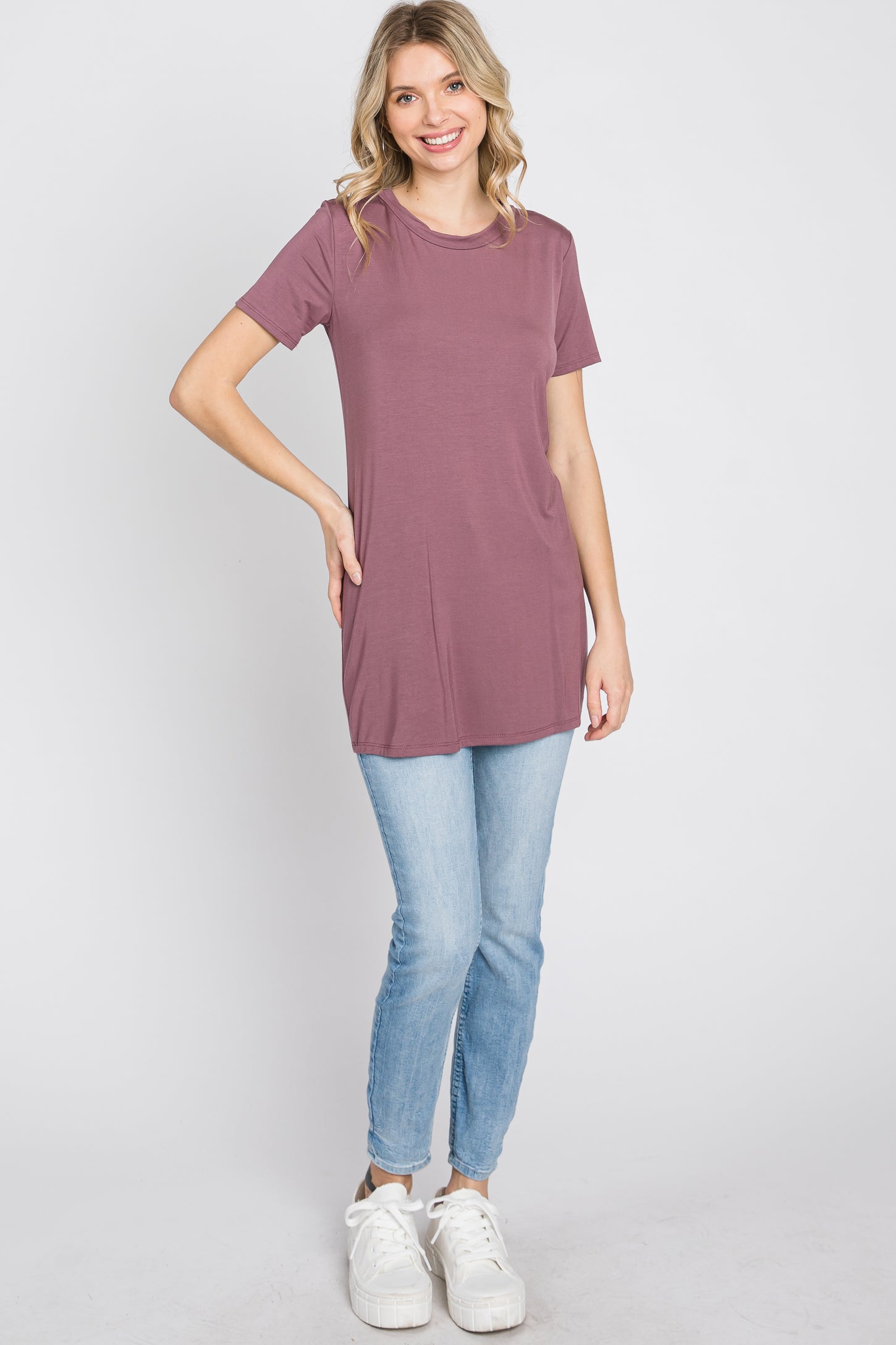 woman wearing a mauve short sleeve tunic top and light blue skinny jeans, posing with one hand on her hip