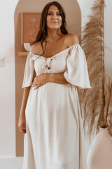woman wearing a beige off-the-shoulder maternity dress with a tie front and flowing sleeves, holding her baby bump