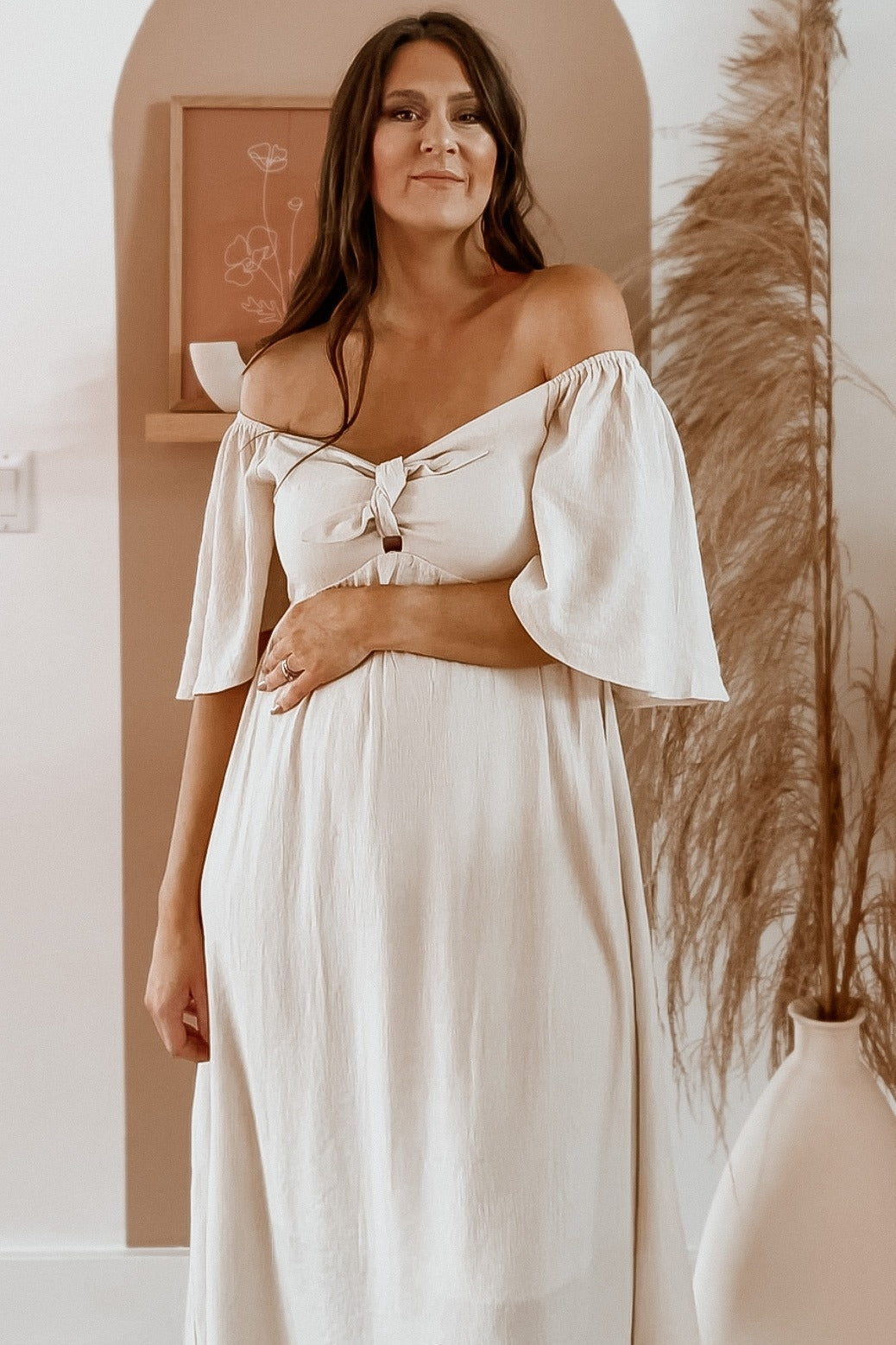 woman wearing a light beige off-the-shoulder maternity dress with a front tie and short flared sleeves, holding her baby bump