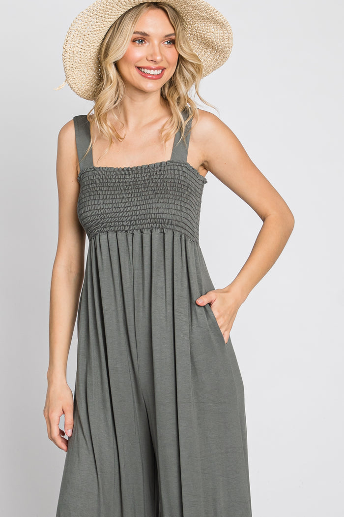 woman wearing a gray smocked jumpsuit with wide legs and pockets, holding a straw hat