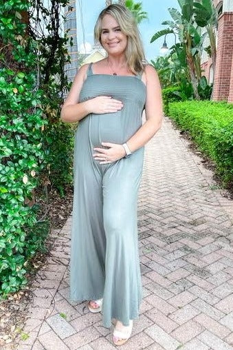 woman wearing a light green maternity jumpsuit and holding her baby bump while standing outdoors.