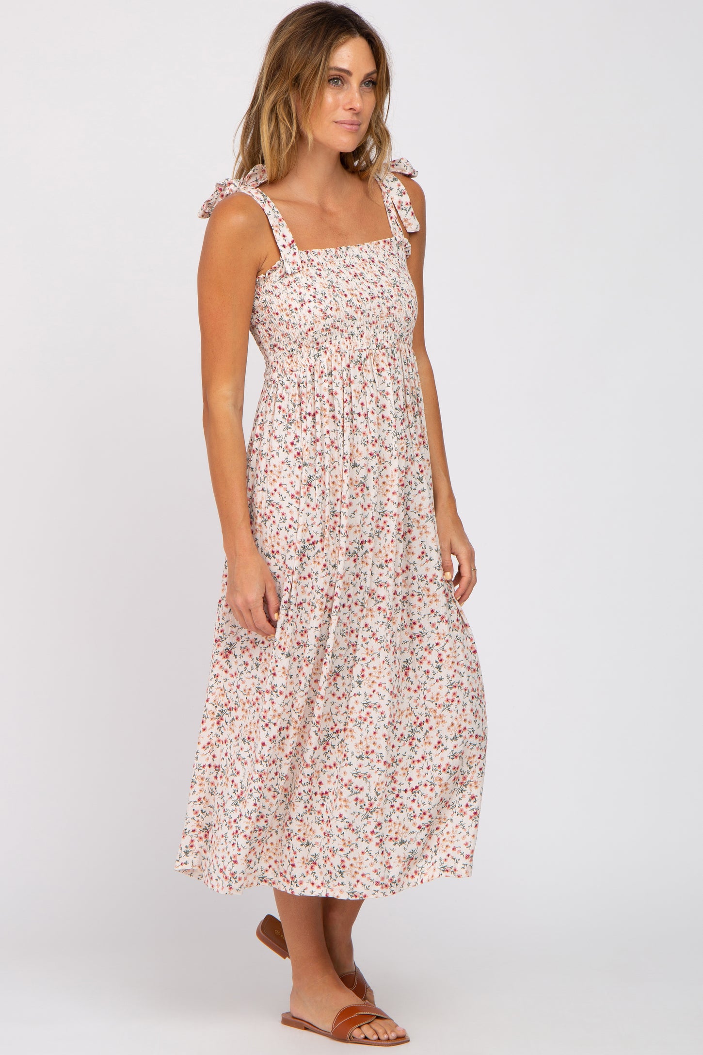 woman wearing a midi-length floral dress with tied shoulder straps and a smocked bodice, paired with tan slide sandals