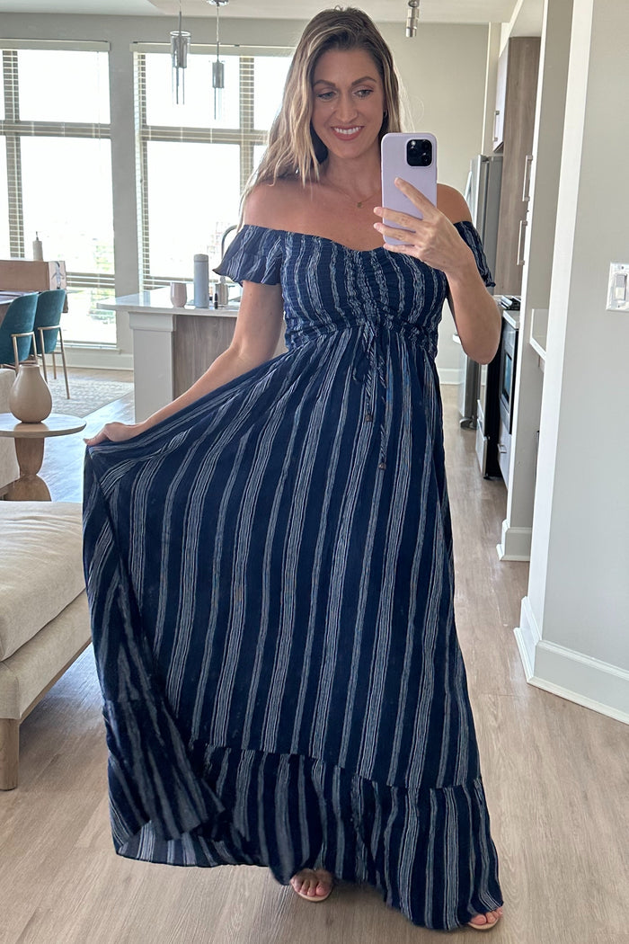 woman wearing a navy blue off-the-shoulder striped maxi dress with a tied waist and ruffled hem