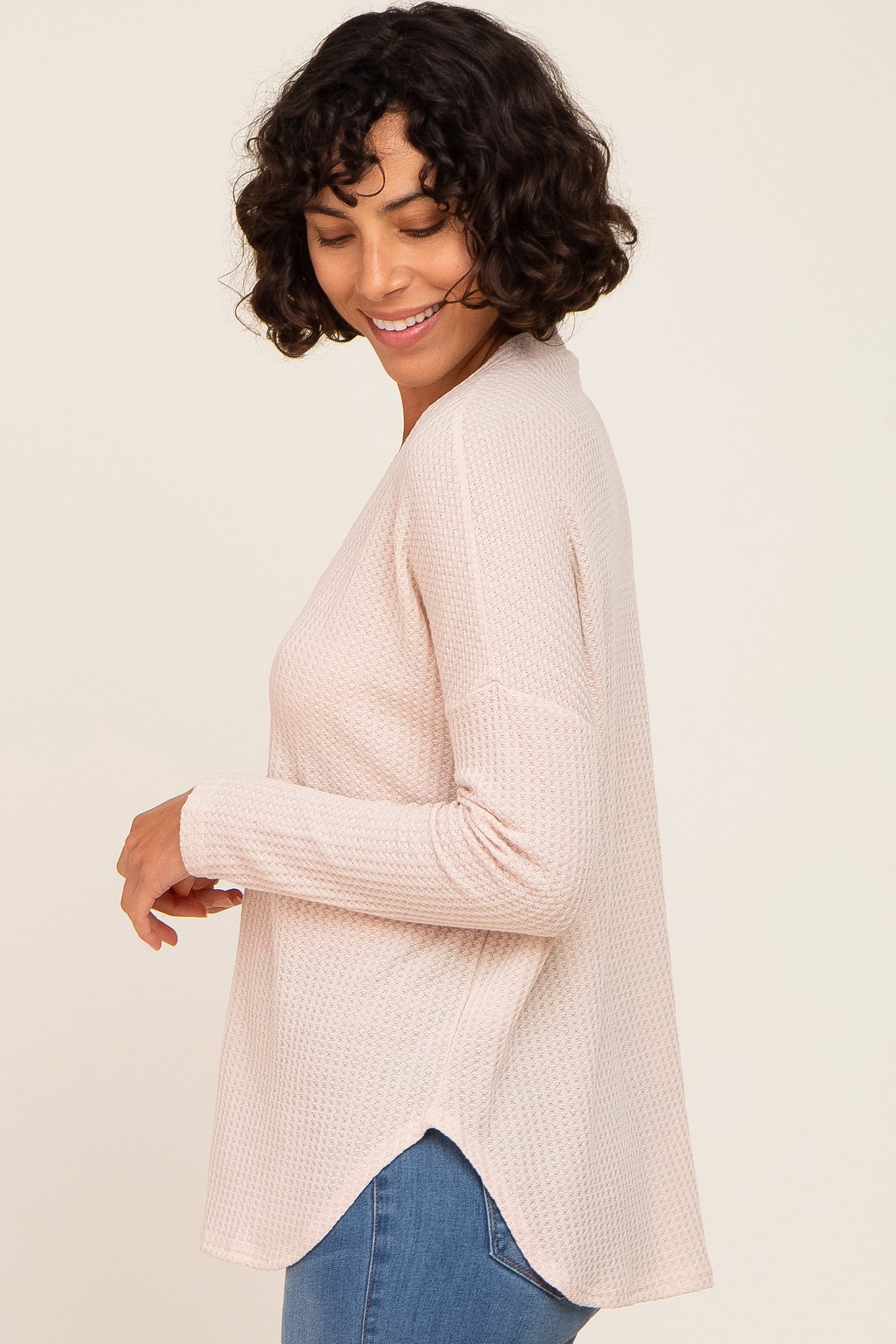 woman wearing a light pink waffle knit long sleeve top with a rounded hem and side slits, smiling and looking down
