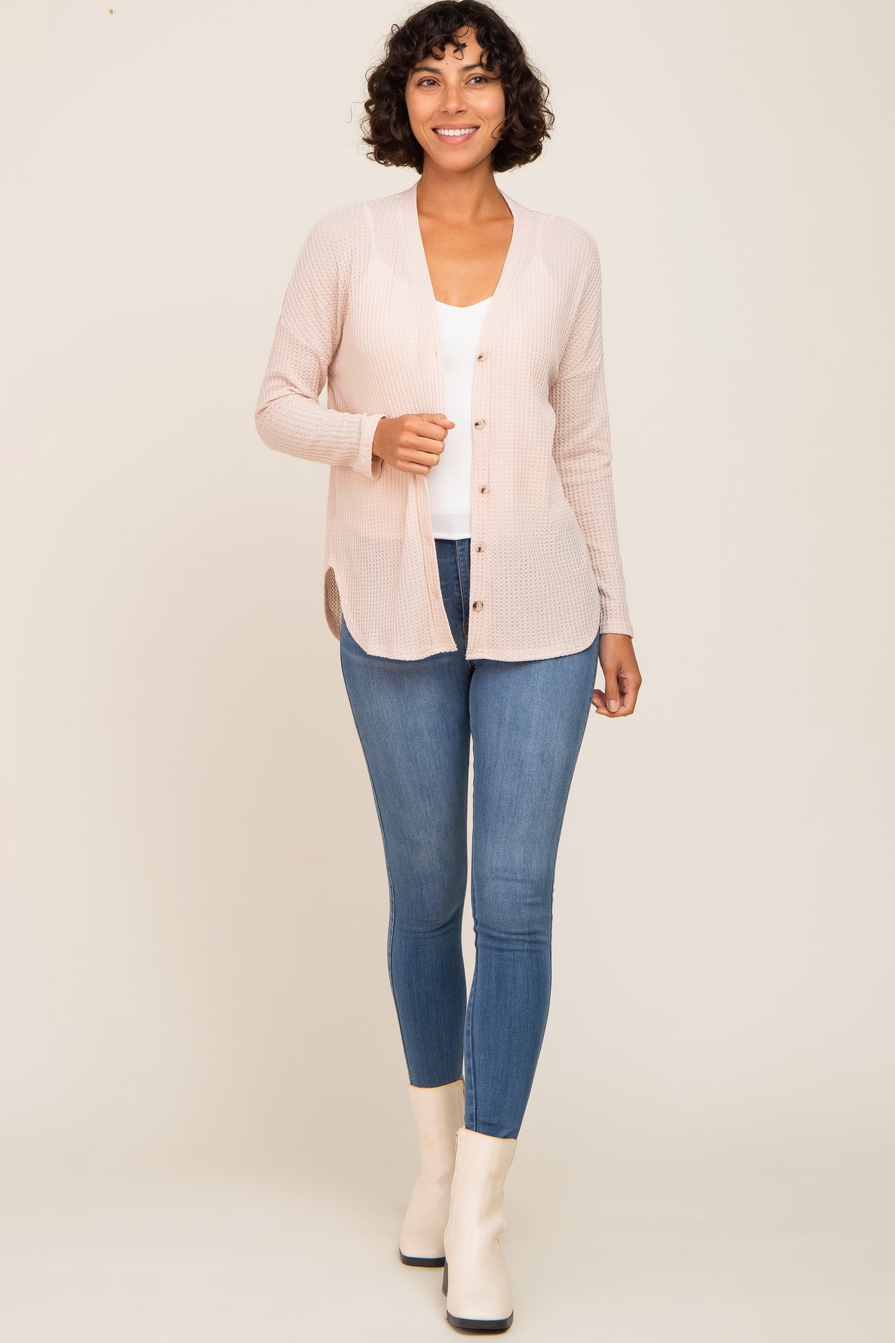 woman wearing a light pink button-up cardigan over a white top and blue skinny jeans, standing and smiling