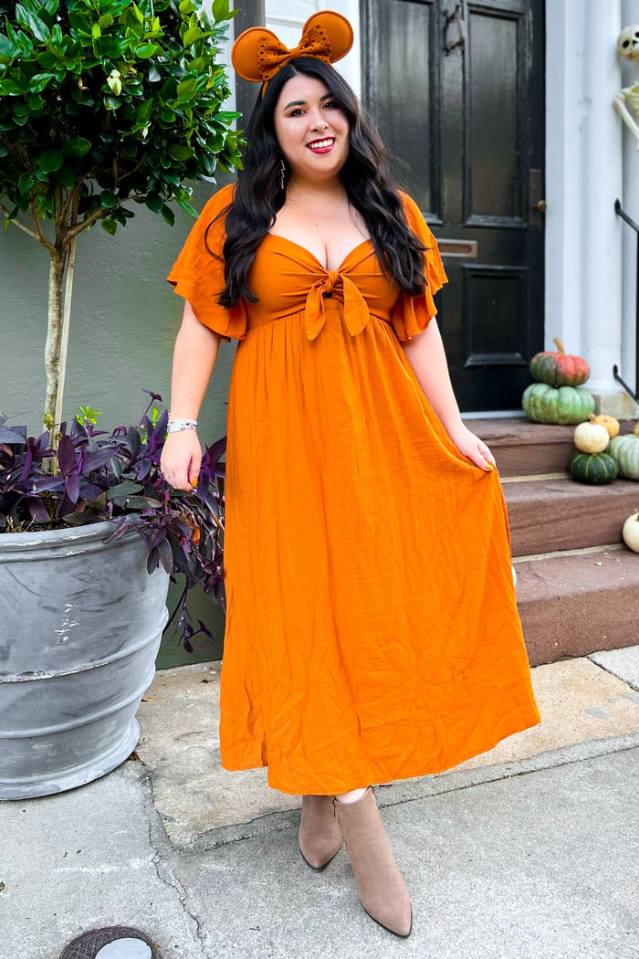 woman wearing an orange maxi dress with flutter sleeves and a tie front, accessorized with mouse ears headband