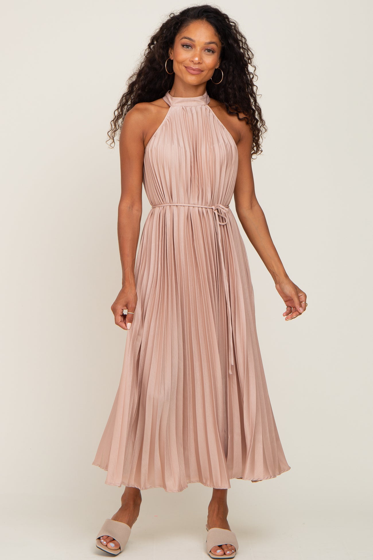 woman wearing a blush pink pleated halter neck dress with a belted waist and curly hair