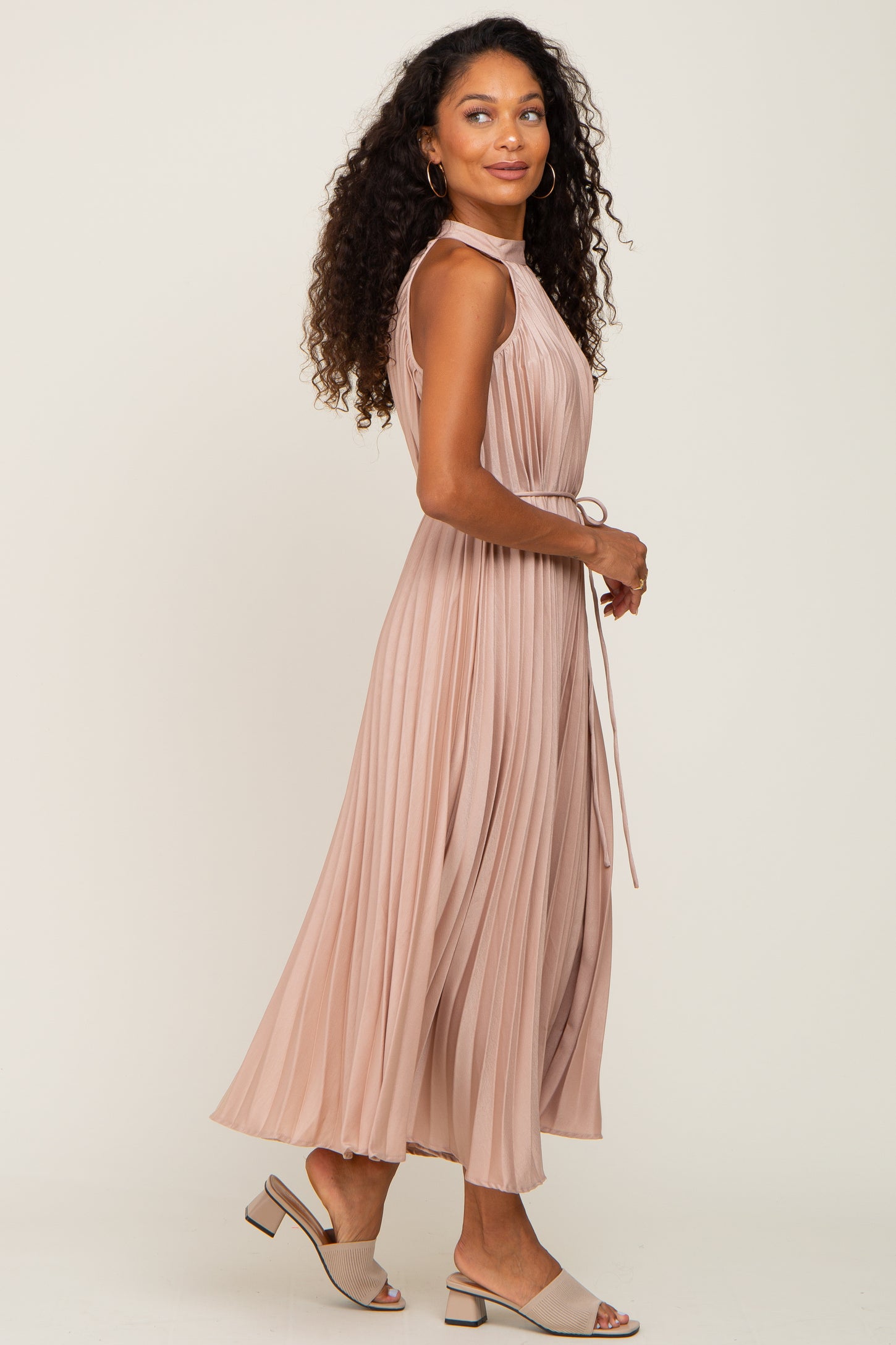 woman wearing a blush pink pleated midi dress with a tie waist and side view of her curly hair.
