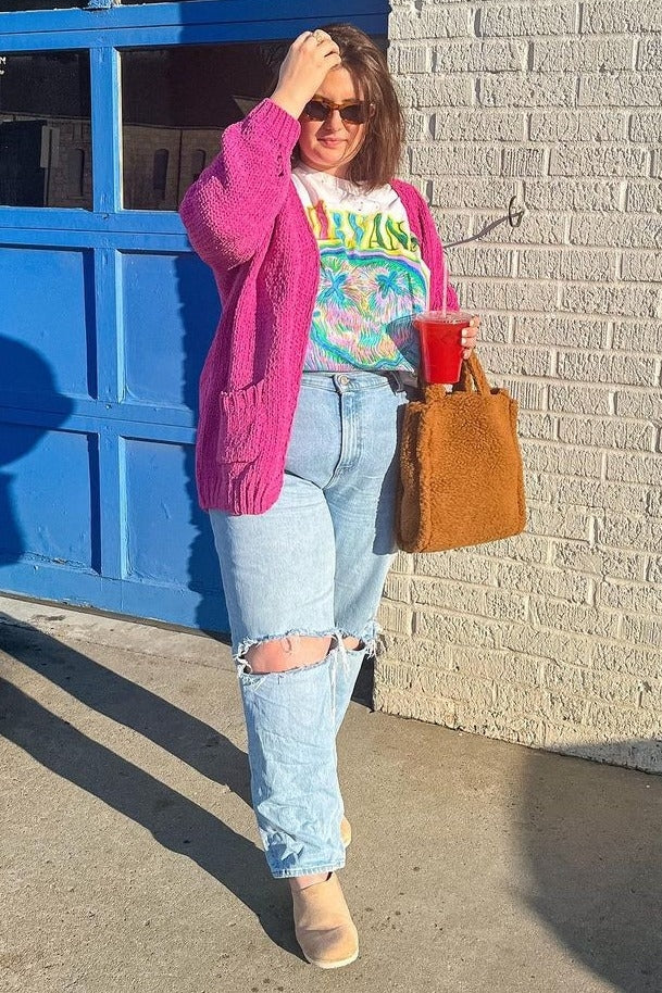 woman wearing a bright pink chunky knit cardigan, colorful graphic t-shirt, ripped light blue jeans, and holding a drink with a brown tote bag