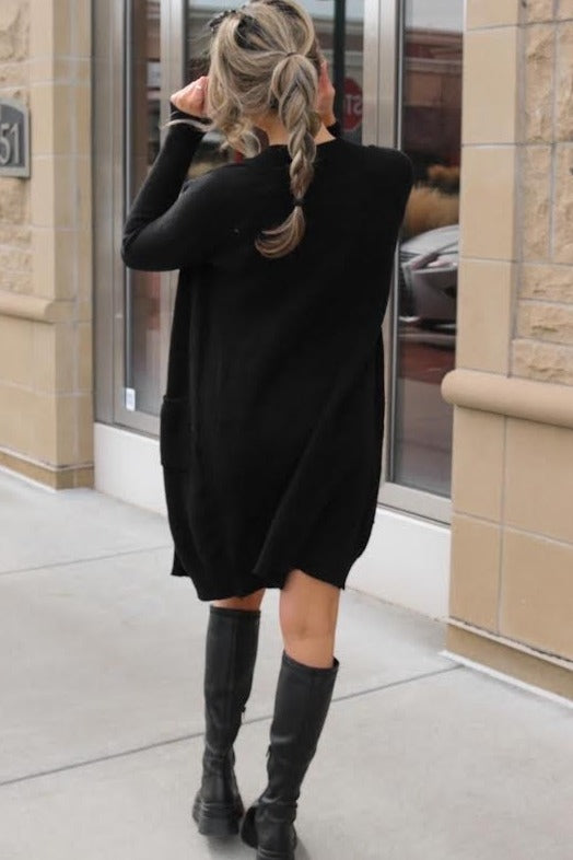 woman wearing a black oversized sweater dress with pockets and tall black boots, viewed from the back