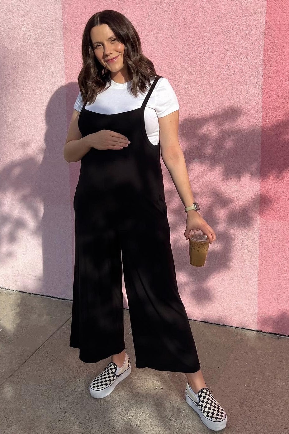 woman wearing a black maternity jumpsuit with a white t-shirt underneath, holding her baby bump and an iced drink