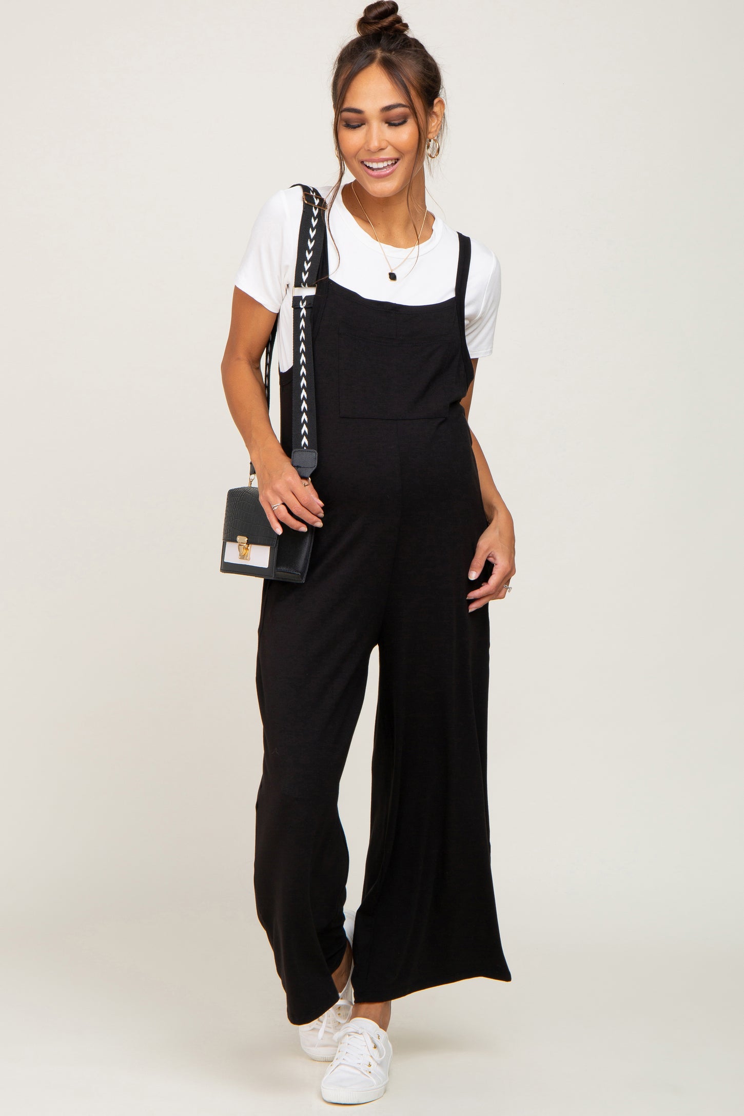 woman wearing a black maternity overall jumpsuit over a white t-shirt, styled with a small handbag and sneakers