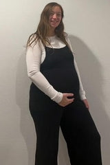 woman wearing a black maternity overall with a white long sleeve shirt, showcasing her baby bump
