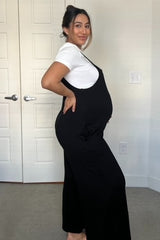 woman wearing a black maternity jumpsuit and a white t-shirt, showing her baby bump while posing in a room