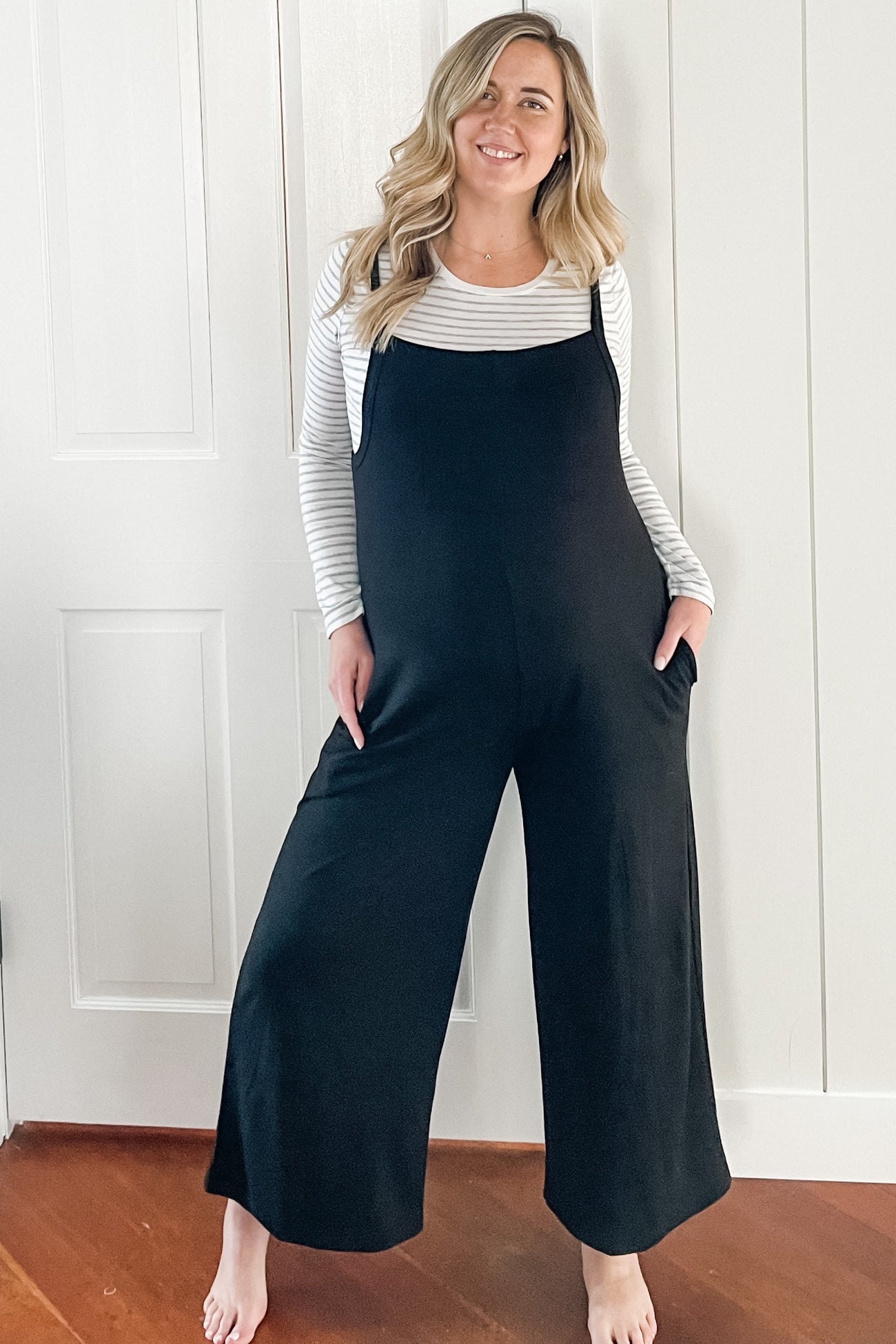 woman wearing a black maternity jumpsuit with wide legs and pockets, paired with a striped long sleeve top