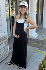 woman wearing a black maternity jumpsuit and white tank top, holding her baby bump while standing outside