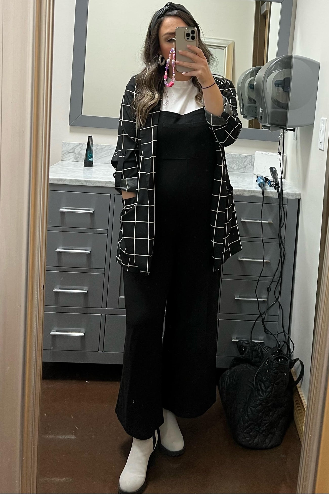 woman wearing a black and white grid patterned maternity shirt over a black maternity jumpsuit and white ankle boots