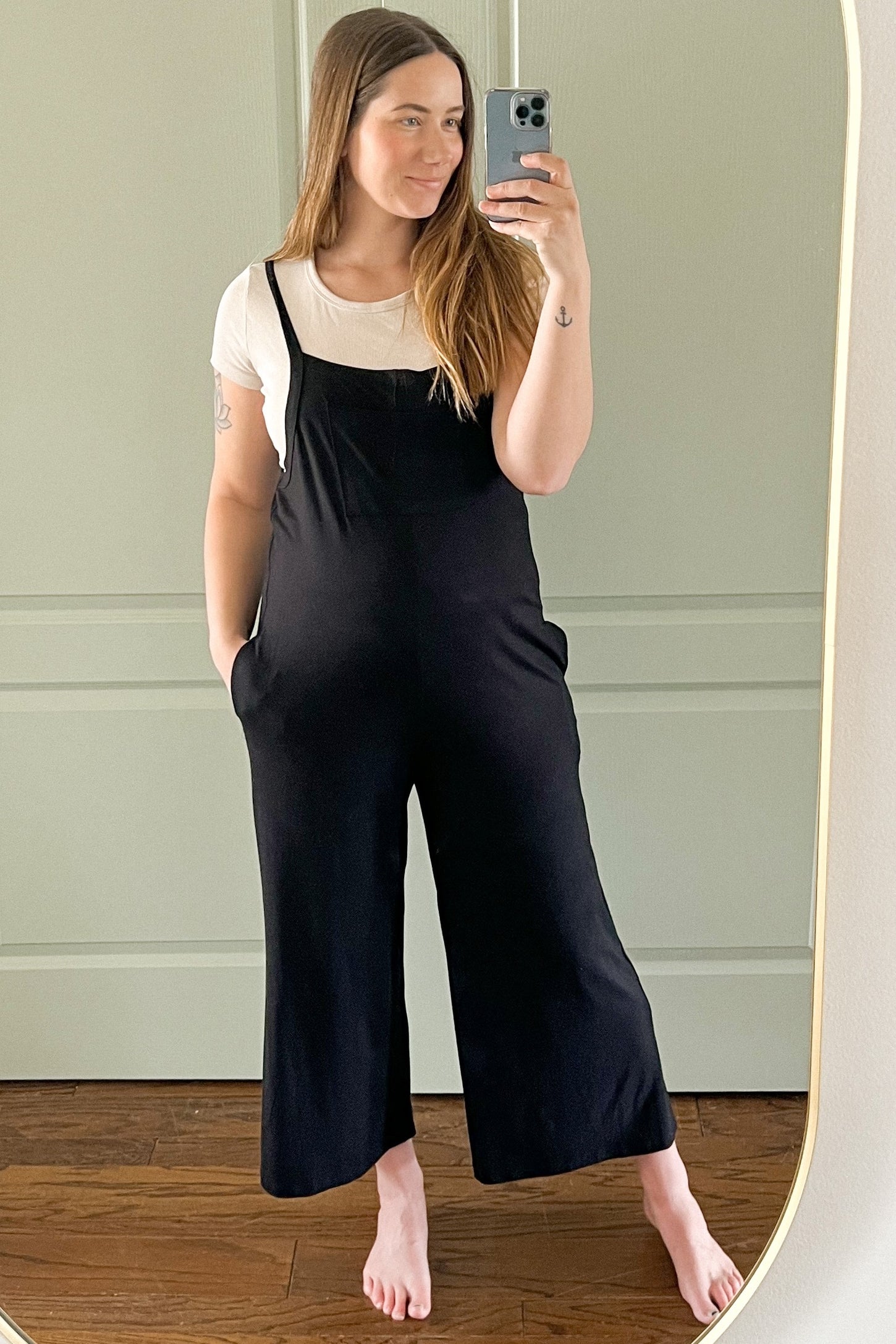 woman wearing a black maternity jumpsuit with wide legs and pockets, posing in front of a mirror with a visible baby bump