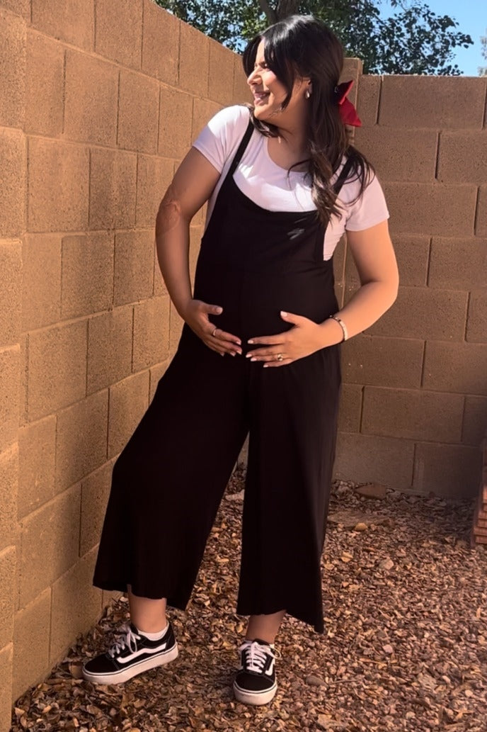 pregnant woman in black maternity overalls and white t-shirt posing against a brick wall