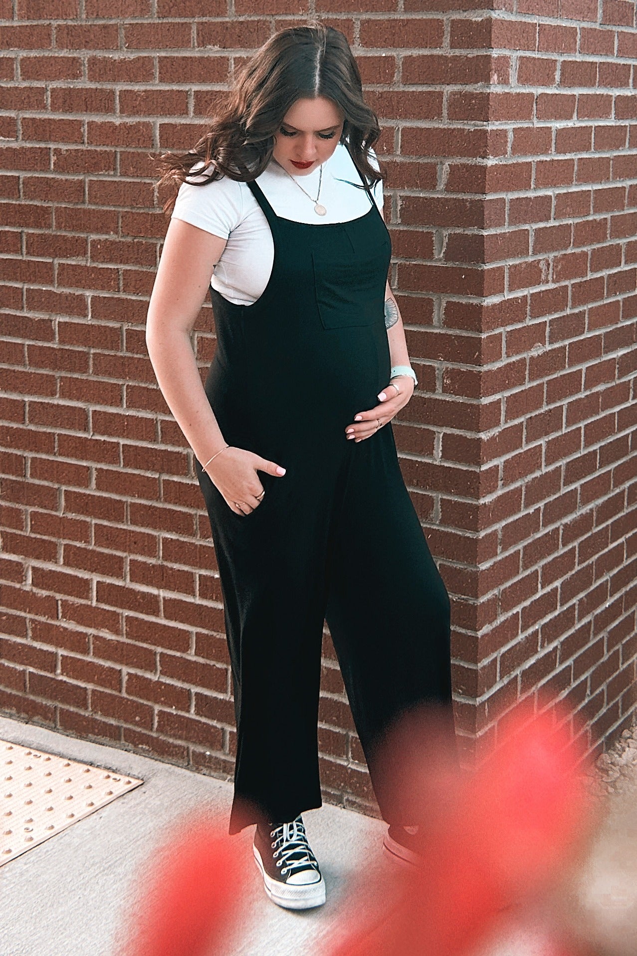 woman wearing a black maternity jumpsuit over a white t-shirt, showing her baby bump while standing against a brick wall
