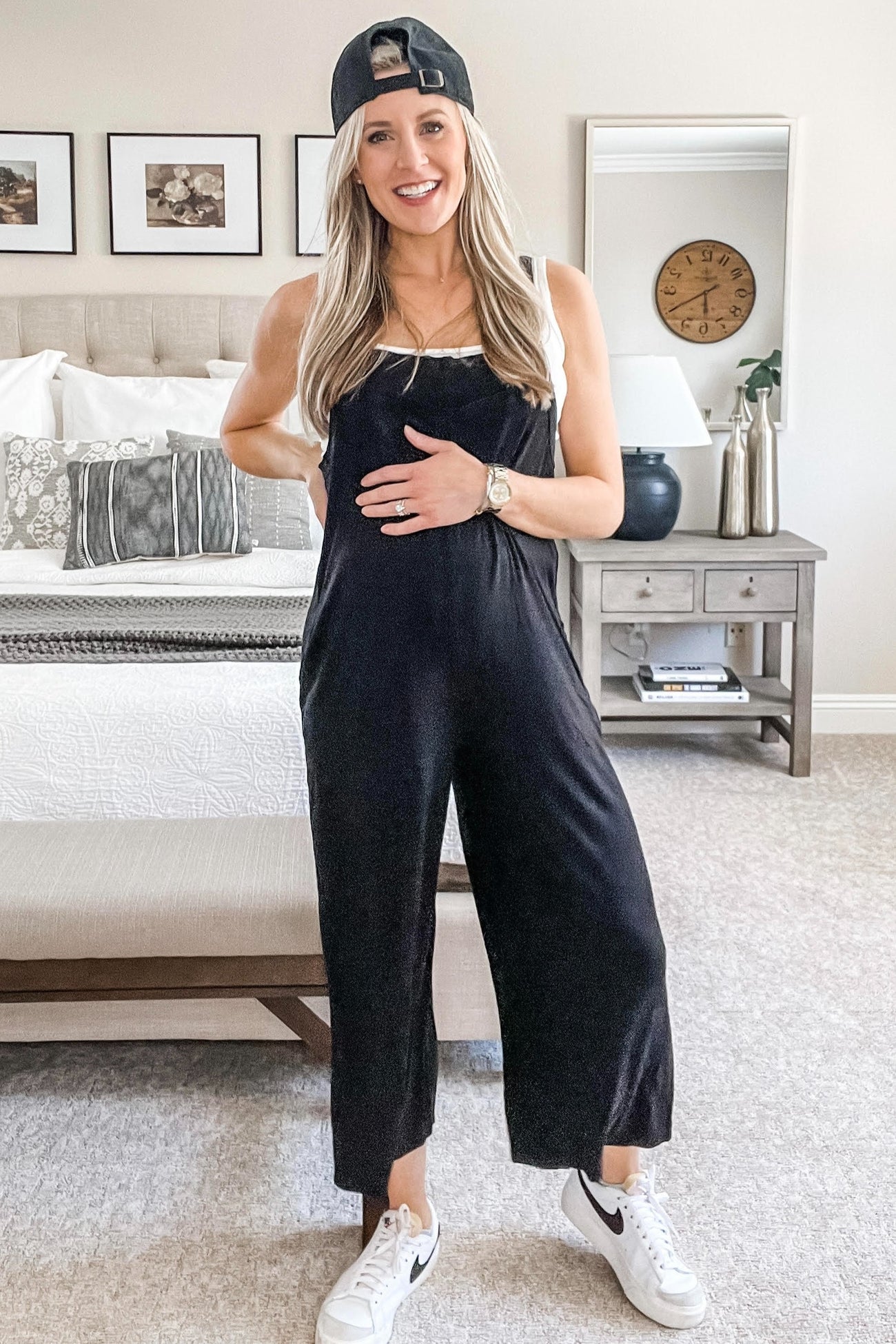 woman wearing a black maternity jumpsuit and holding her baby bump, styled with a cap and white sneakers