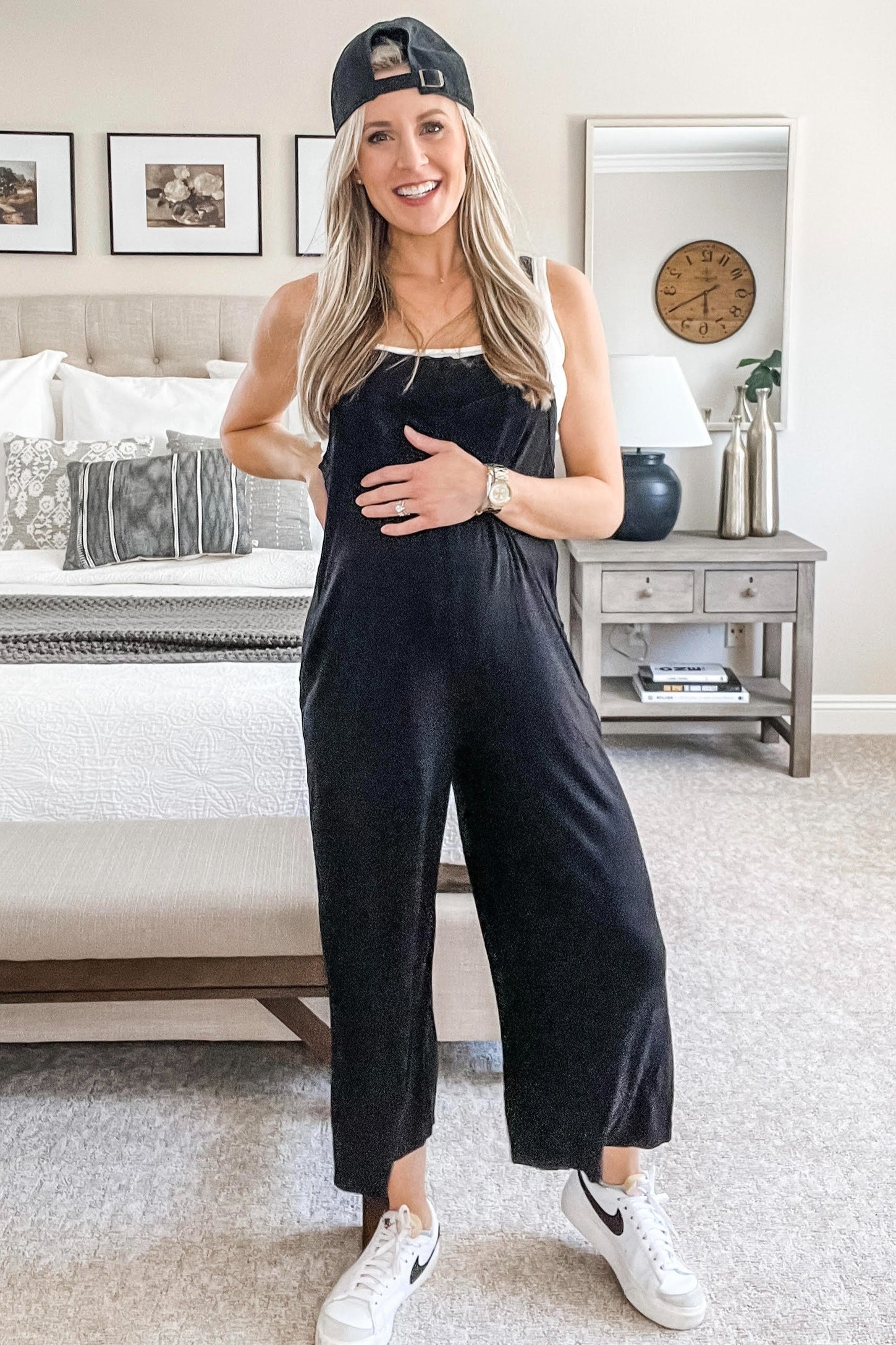 woman wearing a black maternity jumpsuit and holding her baby bump, styled with a cap and white sneakers