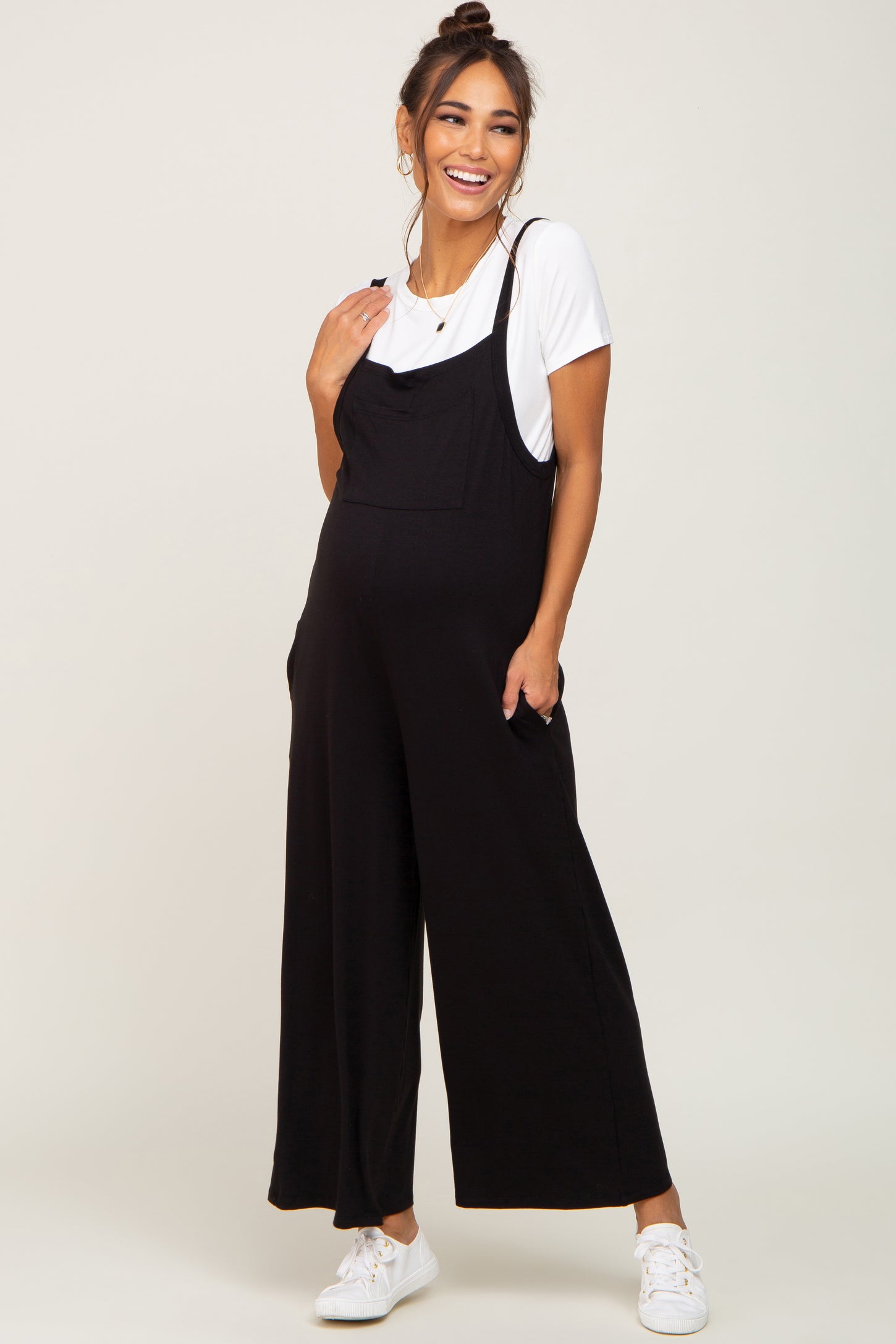woman wearing a black maternity jumpsuit with a white t-shirt underneath and pockets, smiling with a visible baby bump