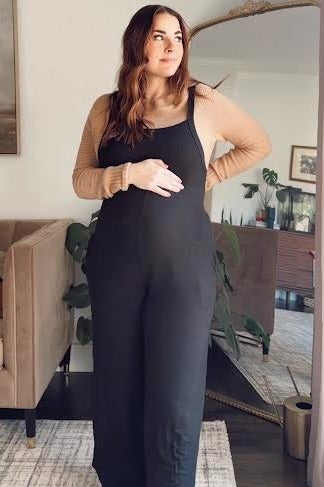woman wearing a black maternity jumpsuit and a beige long sleeve top, gently holding her baby bump